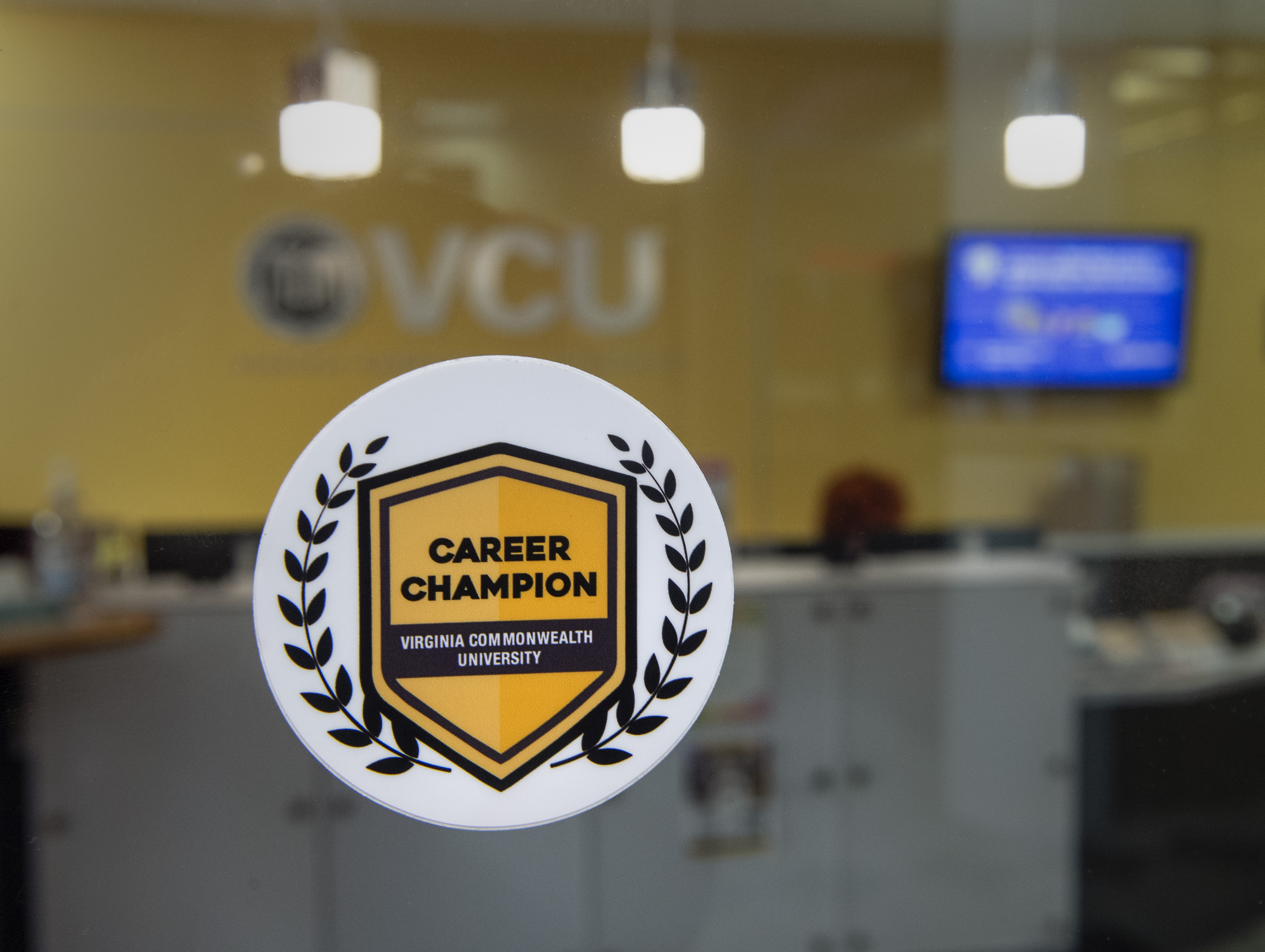 A white sticker with a yellow crest is in the foreground of an office with VCU on the back wall. The sticker reads "Career Champion, Virginia Commonwealth University"