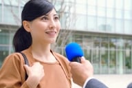 Young Asian woman outside classroom building talking into microphone held by someone off-camera