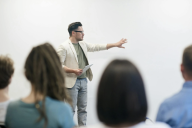 Male professor addresses a group of students before a white background.