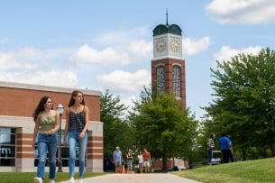Two girls walk on a sidewalk with a college campus behind them. 