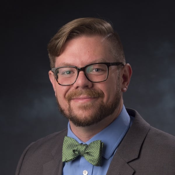 Christopher Haynes, a light-skinned man with brown hair, glasses and a beard who is wearing a blue collared shirt with a green bow tie under a brown jacket.