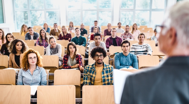 A professor stands in front of a classroom of students with some of the chairs unfilled.  