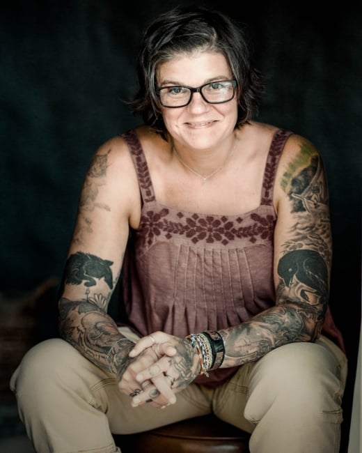A woman in glasses with tattoos sits for a portrait