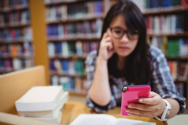 A girl wearing glasses is slightly out of focus and holding a pink phone. She is sitting in a library with bookshelves behind her.