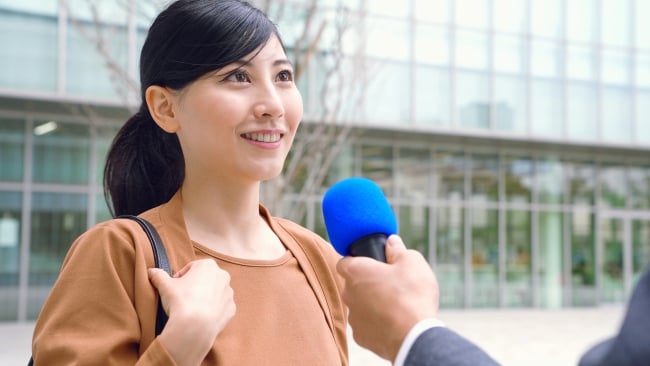 Young Asian woman outside classroom building talking into microphone held by someone off-camera
