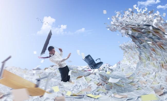 Man stands in a sea of paper holding an umbrella turned inside out as a huge wave of more paper approaches