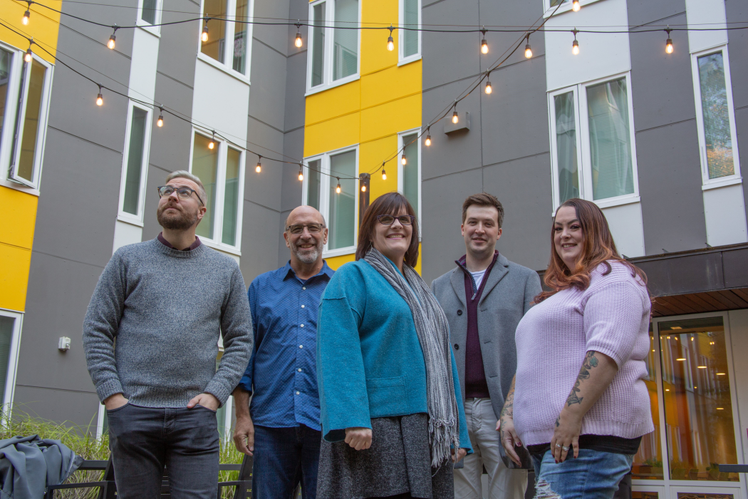 Five of the College Housing Northwest staff smile in front of one of the apartment buildings, which is tiled in gray, white and yellow.