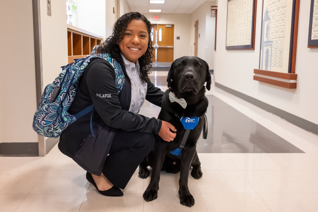Hartwick student Serinah Palafox, a woman with light-brown skin and curly dark hair, smiles with her black Lab puppy in training. The puppy, Stitch, is wearing a bow tie.