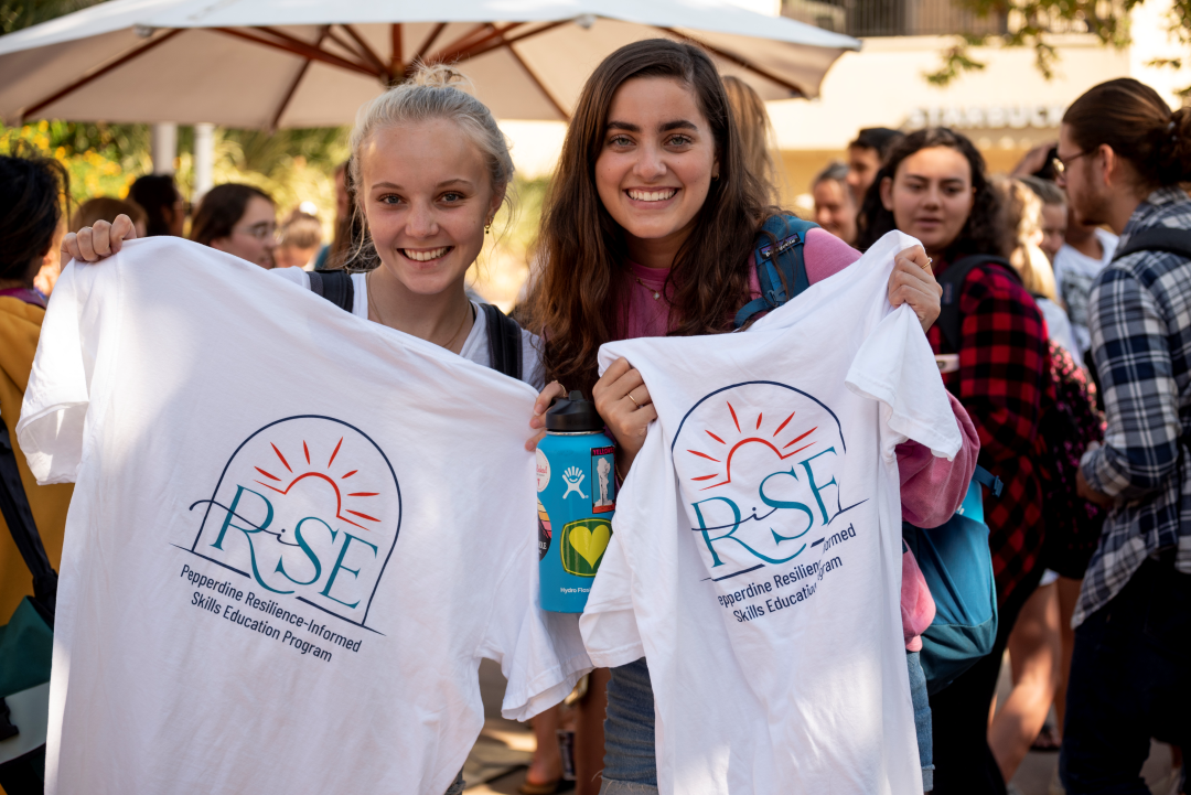Two students smile holding white T-shirts with a RISE logo.