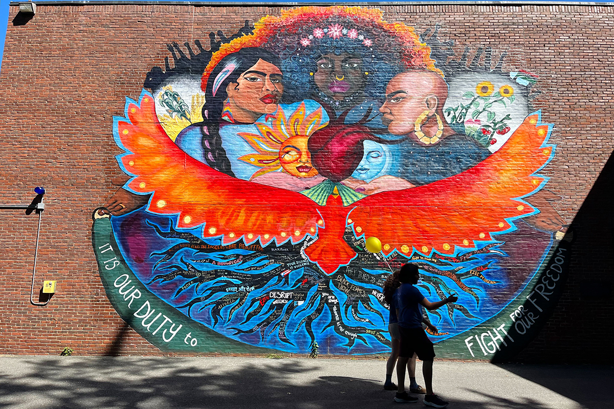 A large mural depicting women of different races atop a large bird. The words at the bottom say, “it is our duty to fight for our freedom.” Two people pass in front of the mural.