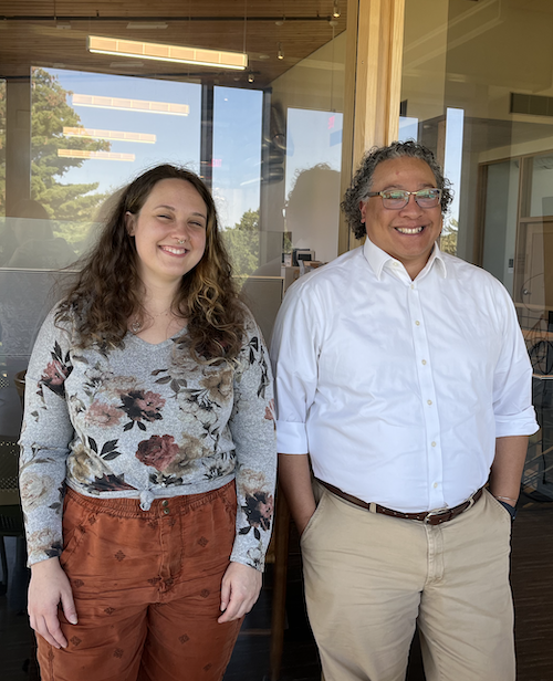 Two people, a woman with light brown curly hair and a man with curly hair and glasses, pose in front of a window.