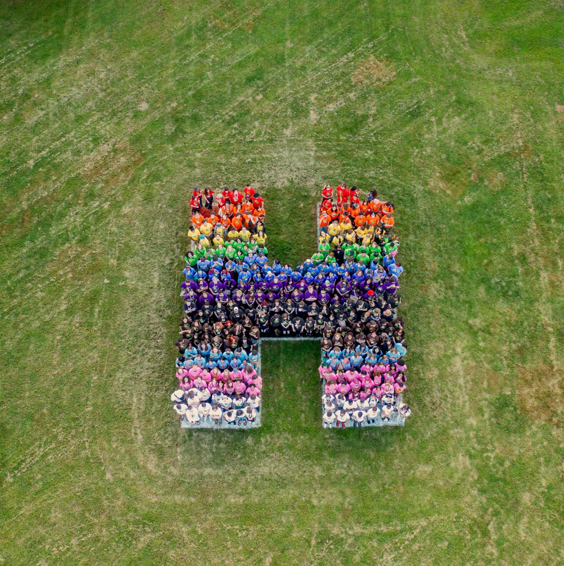 Students form a multicolored H in the colors (from top to bottom) red, orange, yellow, green, blue, purple, black, brown, light blue, pink, white. Behind them is green grass.