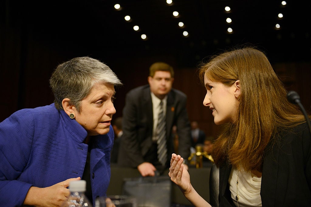 One woman wearing a blue jacket speaks to another woman in a black blazer in a Senate hearing room.