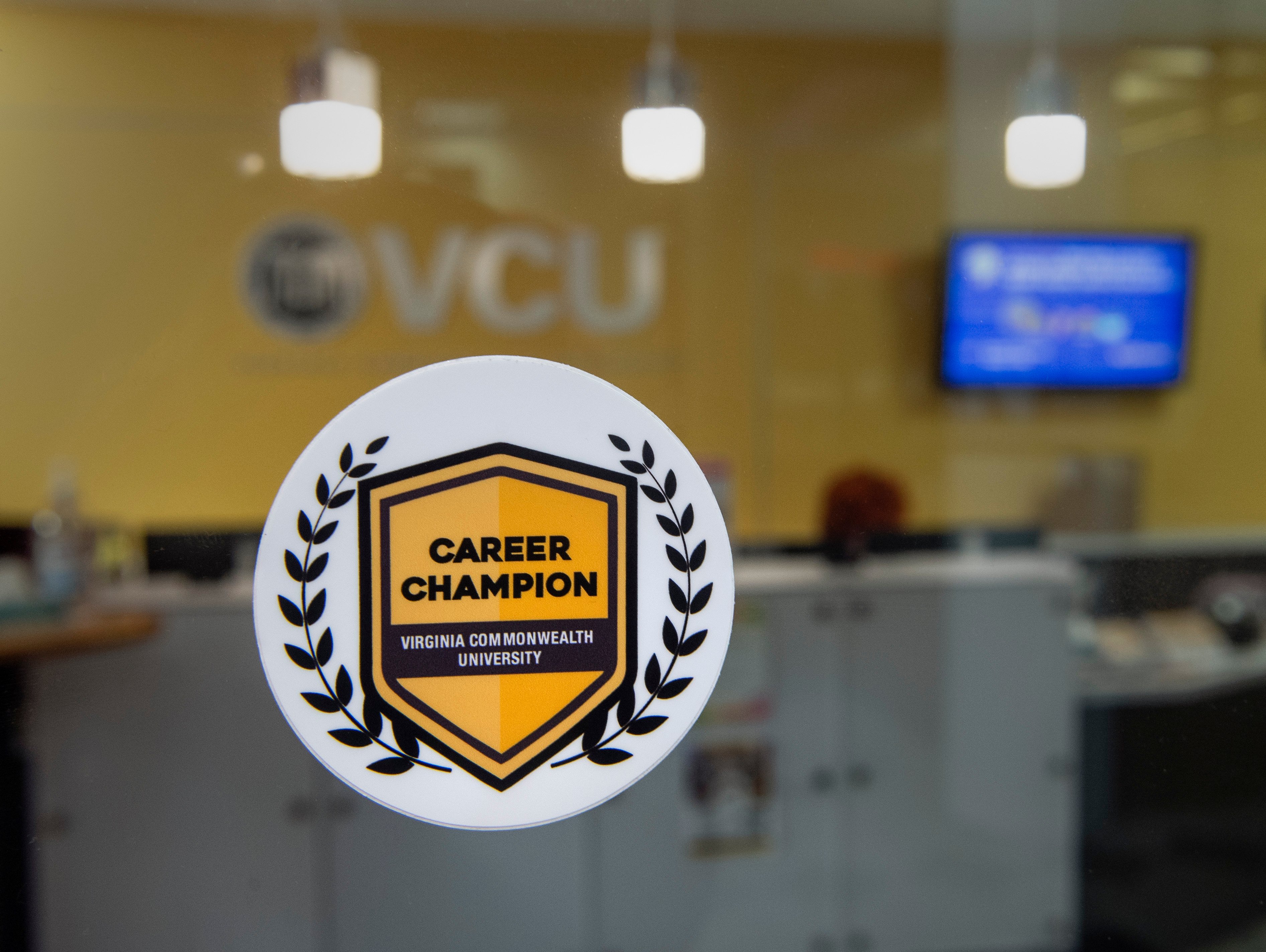 A white sticker with a yellow crest is in the foreground of an office with VCU on the back wall. The sticker reads "Career Champion, Virginia Commonwealth University"