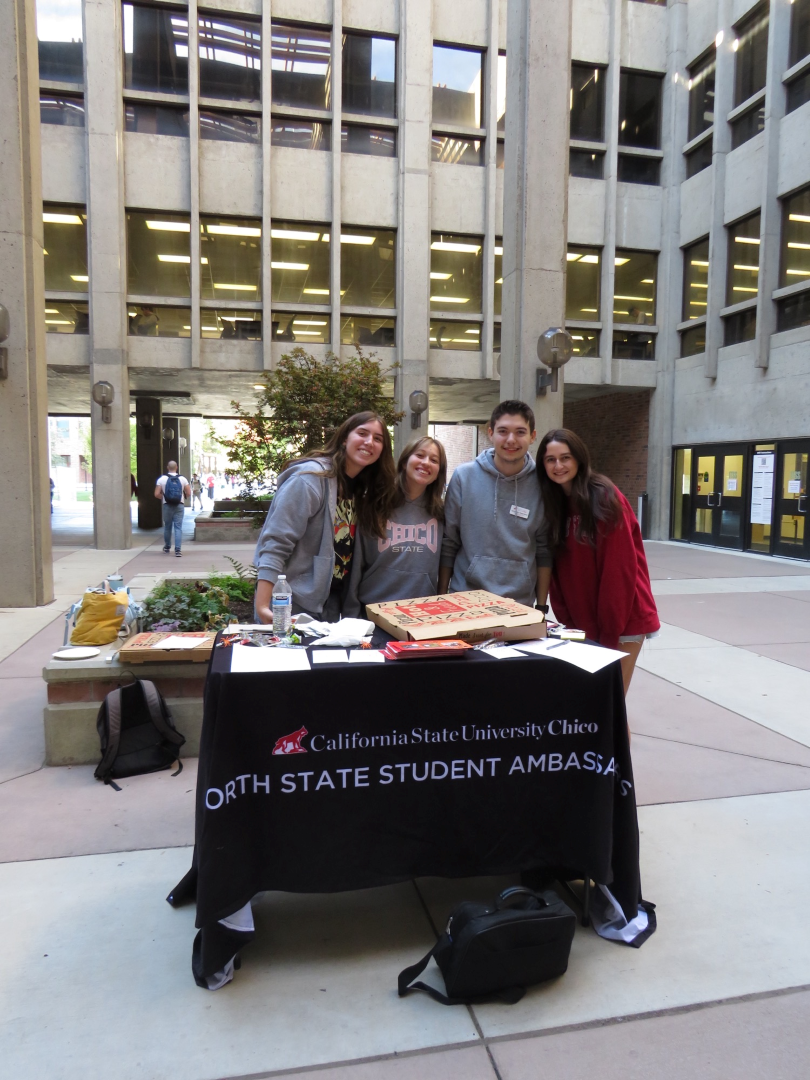 Four students stand behind a table with a black table cloth that says "California State University Chico North State Student Ambassadors." The students are outside on a sunny day and there are pizza boxes on and around the table.
