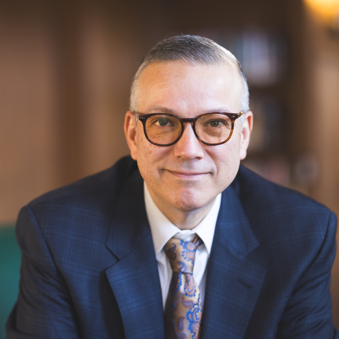 Estevan Garcia smiles for a headshot in a blue suit jacket and paisley tie with brown framed glasses