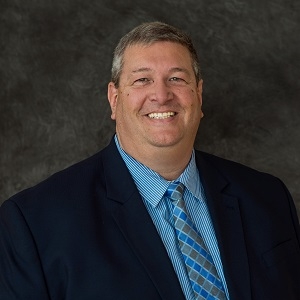 Ron Slinger smiles for a headshot in front of a gray backdrop wearing a blue shirt and tie under a black jacket.