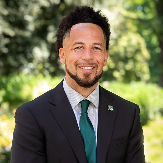 Sacramento State University president Luke Wood smiles for a headshot in a black suit coat, green tie and Sac State pin on his lapel