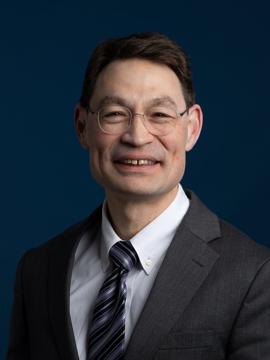 John Su smiles for a headshot wearing thin framed glasses, a white shirt, black suit coat and dark patterned tie