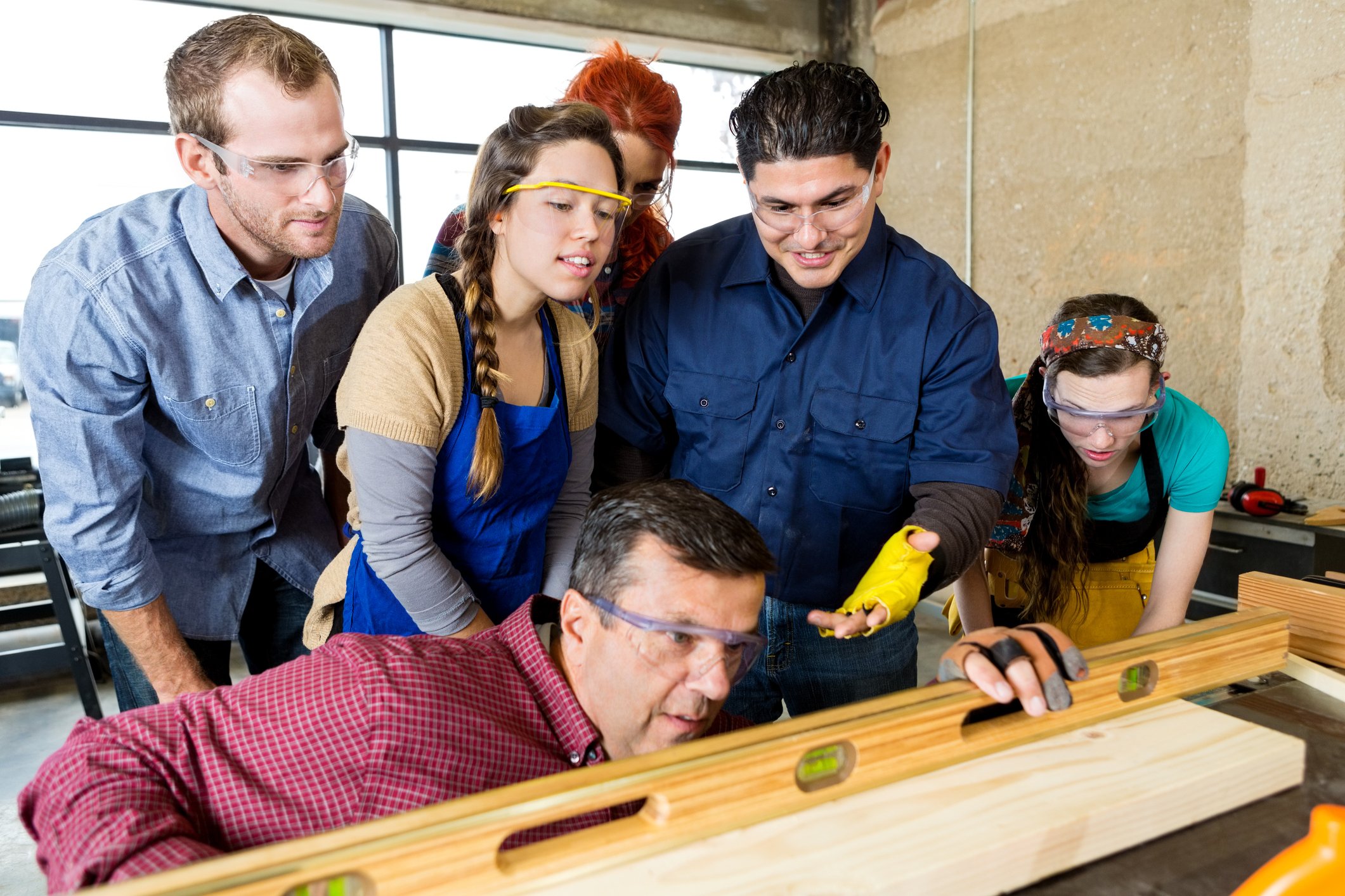 Diverse group of woodworking workshop employees watch as their manager uses a leveler tool on wood board. The employees are attentively watching and a man asks the manager questions. Large bright windows are in the background.