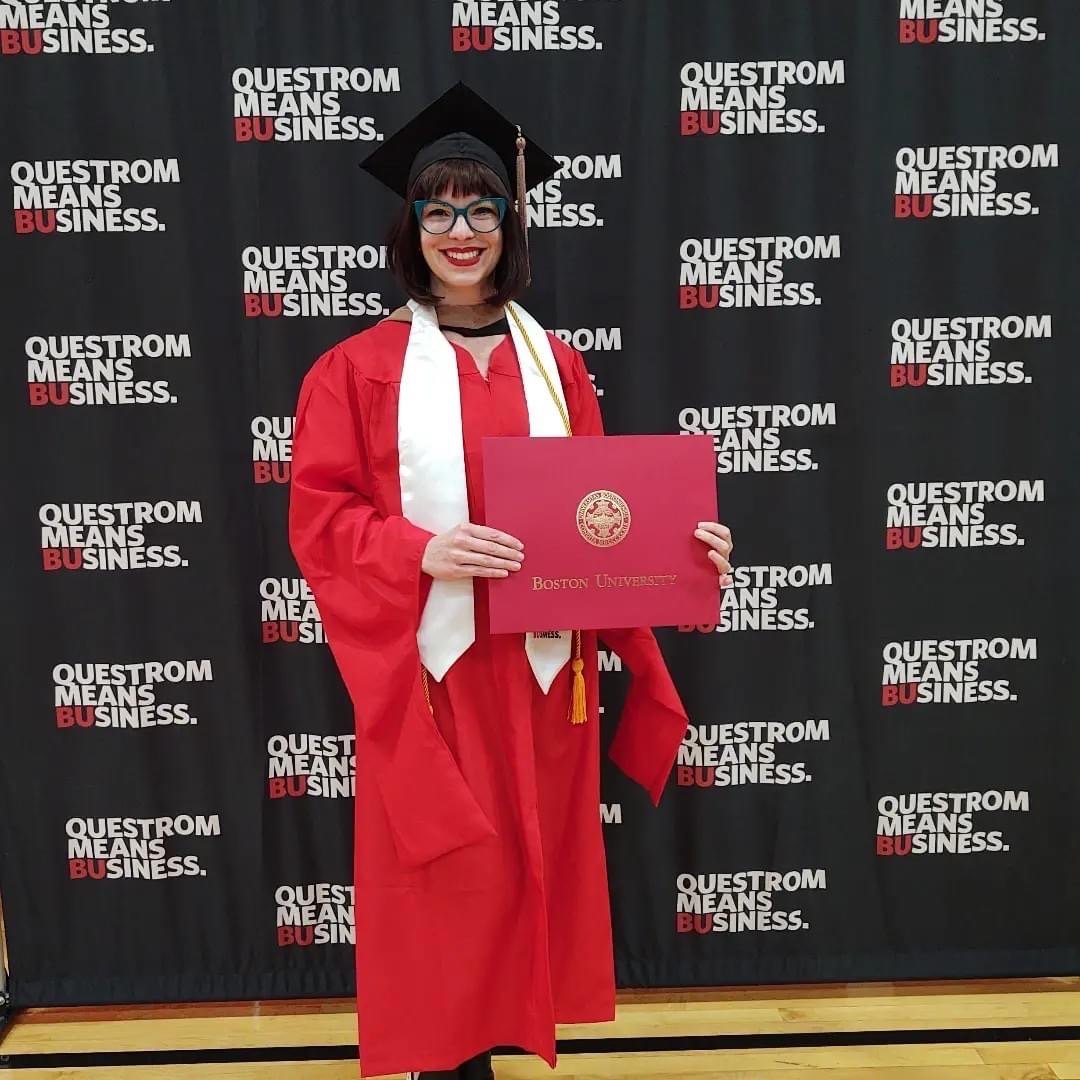 A light-skinned woman with dark hair and glasses wearing academic regalia and holding her diploma.