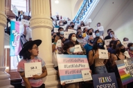 Protesters inside a building holding signs opposed to a ban on transgender student athletes.