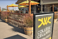 Two people sit in the background of a winery patio under a yellow umbrella. Sign in front of frame says College Cellars.