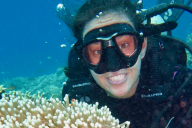 In an underwater photo, a woman in a scuba mask smiles while next to a coral reef