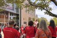 People, mostly in red shirts, some with #campusworkers4democracy on the back, listen to a speaker with a megaphone outside a Virginia Commonwealth University building.