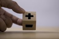 A wooden cube has a subtraction sign on one face and a subtraction sign on another. A hand flips the cube away from the addition sign in favor of the subtraction sign.