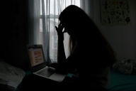 Young woman sits in a dark room in silhouette looking at her open laptop. 