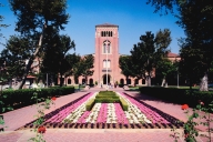 A University of Southern California campus photo, depicting a long walkway toward the entrance of a USC building. A garden with pink and white flowers and well-manicured shrubbery bisects the brick walkway. 
