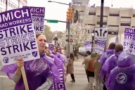 People, most wearing purple ponchos, hold purple and white signs on a sidewalk. The signs say "UMich Grad Workers Strike Strike Strike" and the union's name.