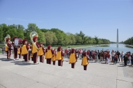 A marching band in red and gold outfits plays in front of the Reflecting Pool and Washington Monument.