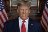 Donald Trump, wearing a blue suit and red tie, speaks in front of two American flags.