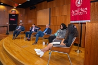 On a large wooden stage, four trustees and a student sit in wooden chairs facing an audience for a Q&A-style discussion.