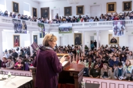 A woman with her back to the camera stands at a lectern, addressing a room full of students. The room appears to be a historic building, with seating on the floor and up in a gallery.