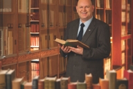Alan Bearman, a bald man with light skin wearing a business suit, smiles holding a book in a library.
