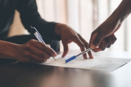 A pair of hands with dark skin works on filling out a form with a pen while another hand with dark skin points at the form with another pen.