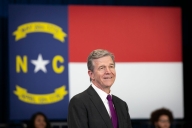 A light-skinned man in a suit and tie with gray hair stands in front of the North Carolina state flag