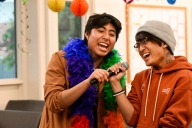 Two students sing karaoke surrounded by rainbow decorations, one of them wearing a rainbow boa