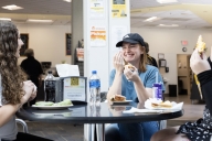 Three female students sit at a small table in a cafeteria eating sandwiches, laughing. 