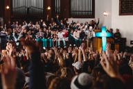 A crowd of students and visitors hold their hands up at Asbury University's chapel as someone preaches from a podium with a large blue cross. 