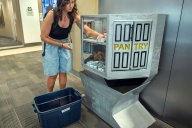 A student wearing a backpack helps restock a gray food pantry from a blue plastic tub.