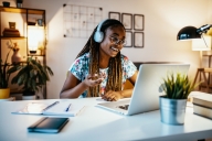 An African American student talks with peers online wearing headphones at her desk.