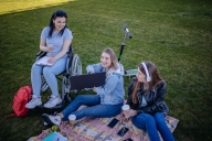 Three female college students sit outside on the grass on a blanket, laughing and looking at a computer screen. One of the women is using a wheelchair.