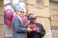 Two people stand together holding a maroon Aggie shirt with maroon and silver balloons behind them. 