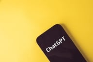 A black mobile phone with the words "ChatGPT" in bold white letters on the screen, against a yellow background.