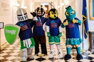 Four mascots—a knight, a badger, a hornet and a hawk—pose next to one another in identical shirts in a college building.