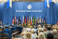 Commencement at Dickinson State University, with blue-robed graduates sitting on the left side of the photo and an array of flags up on the dais behind a speaker in academic regalia.